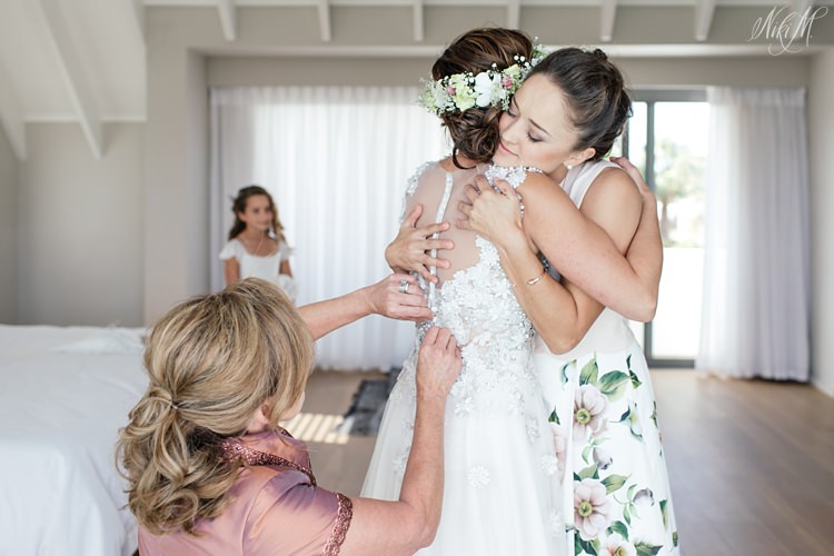 Bride and sister embrace whilst the mother of the bride buttons up the wedding gown