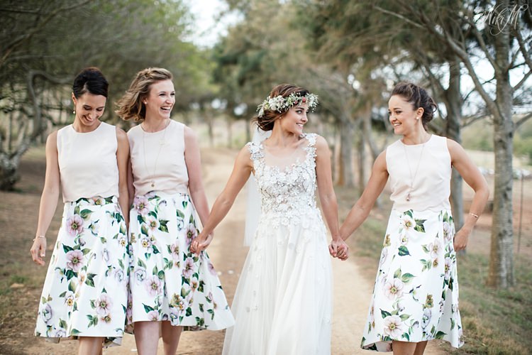 Bride and bridesmaids in floral print Jenni Button dresses walk down a farm road laughing